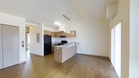 This single-floor space showcases a clean, modern design with an open-concept layout that seamlessly connects the kitchen and living area. The kitchen is a standout feature, equipped with warm wood cabinetry, granite-patterned countertops, and black appliances including a full-size refrigerator and range. A peninsula counter extends into the living space, offering additional prep area and casual seating potential, while a trio of sleek pendant lights hang above, adding a stylish focal point to the room. Recessed and flush-mounted lighting provides bright, even illumination throughout. The flooring is a rich hardwood-style material that runs continuously across the open space, lending warmth and cohesion to the interior. The vaulted or high ceilings amplify the sense of openness and airiness, making the space feel generously proportioned. Crisp white walls serve as a neutral backdrop, allowing the natural wood tones of the cabinetry and flooring to stand out. An electrical panel is accessible from the main area, and the overall layout is thoughtfully designed for functionality and ease of movement. This space offers a welcoming, contemporary atmosphere well-suited for comfortable everyday living.