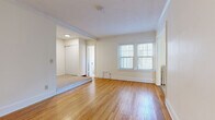 This single-floor unit offers an efficiently organized layout comprising a living room, bathroom, and laundry area. The living room is the largest space, characterized by warm-toned hardwood floors that run throughout and crisp white walls accented by crown molding. Double windows allow natural light to fill the room, contributing to a bright, open feel despite the unit's compact footprint. A stepped transition leads to a carpeted alcove area, which provides additional functional space and includes a closet for storage. A wall-mounted radiator is also present, indicating a classic heating system.

The bathroom maintains a consistent, clean aesthetic with white subway tile covering the walls, a mosaic tile floor, and a wall-mounted pedestal sink with chrome fixtures. A soaking tub sits beneath a window fitted with blinds, and a mirrored medicine cabinet completes the space.

The laundry area accommodates a stacked washer and dryer unit, offering in-unit convenience. Throughout the space, white baseboards and neutral wall tones create a cohesive, understated palette. The overall design reflects classic architectural detailing, with a straightforward and functional arrangement suited for efficient single-occupant living.