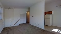 This single-floor space features a modest, open layout with neutral white walls and wall-to-wall carpeting throughout the main living area, creating a clean and functional environment. The living space is straightforward in design, offering a blank canvas for personalization. A built-in closet with classic bifold doors provides convenient storage along one wall, while a hallway beyond leads further into the unit, connecting the living area to additional spaces. Steps near the closet suggest a subtle change in floor level, adding a slight architectural detail to the layout. Adjacent to the living area, the kitchen is partially visible and includes wood-toned cabinetry and a white refrigerator, offering practical cooking and storage functionality. The overall aesthetic is simple and utilitarian, with white painted walls, standard trim, and ceiling-mounted lighting providing adequate illumination. The space is well-suited for those seeking a straightforward, easy-to-maintain living environment with the essentials already in place.