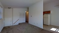 This single-floor space features a modest, open layout with neutral white walls and wall-to-wall carpeting throughout the main living area, creating a clean and functional environment. The living space is straightforward in design, offering a blank canvas for personalization. A built-in closet with classic bifold doors provides convenient storage along one wall, while a hallway beyond leads further into the unit, connecting the living area to additional spaces. Steps near the closet suggest a subtle change in floor level, adding a slight architectural detail to the layout. Adjacent to the living area, the kitchen is partially visible and includes wood-toned cabinetry and a white refrigerator, offering practical cooking and storage functionality. The overall aesthetic is simple and utilitarian, with white painted walls, standard trim, and ceiling-mounted lighting providing adequate illumination. The space is well-suited for those seeking a straightforward, easy-to-maintain living environment with the essentials already in place.