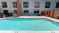 This outdoor pool area combines modern amenities with industrial architectural character to create an inviting and stylish retreat. The centerpiece is a generously sized swimming pool with clear, turquoise water, surrounded by a spacious tiled deck that provides ample room for relaxation and movement. A row of teal-colored lounge chairs lines the poolside, offering comfortable spots to unwind, while a coordinating teal umbrella provides shaded seating nearby. Thoughtfully placed wooden privacy screens and lush landscaping with low-growing shrubs and greenery add warmth and natural texture to the space. The surrounding building façade is a compelling mix of white-painted masonry and exposed brick, with large, black-framed industrial-style windows that lend the area a distinctive urban character. A secondary seating area with outdoor furniture is visible in the background, offering additional space for gathering and socializing. The overall ambiance is energetic yet relaxed, blending the clean lines of contemporary outdoor design with the raw aesthetic of a converted industrial building, making it an ideal space for leisure and recreation.