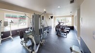 This fitness center is a fully outfitted exercise facility designed to support a well-rounded workout routine. The space is finished with durable dark rubber flooring throughout, providing a safe and functional surface for exercise. Natural light floods the room through multiple large windows, offering views of the surrounding greenery and creating an open, energizing atmosphere complemented by recessed ceiling lighting.

The facility is equipped with a multi-station cable strength machine, ideal for a wide variety of resistance training exercises. Cardio equipment includes elliptical trainers, a stationary spin bike, a treadmill, and a rowing machine, providing ample options for aerobic conditioning. A wall-mounted television is positioned for easy viewing during workouts, and a dry-erase board is available for tracking routines or posting notices.

Full-length mirrors line one wall, enhancing the sense of space while allowing users to monitor their form. The center is accessed through a glass-paneled entry door, with a clearly posted rules and hours notice near the entrance. The clean, neutral-toned walls and well-organized layout contribute to a functional and focused environment suitable for a variety of fitness levels and training styles.