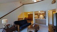 This two-story space is defined by its dramatic vaulted ceilings and open-concept design, creating a sense of generous vertical space in the main living area. A prominent diagonal beam runs across the upper portion of the room, adding architectural character and drawing the eye upward toward the bright, elevated ceiling.

The living area is anchored by a wood-framed futon sofa and a carved wooden coffee table, providing a comfortable and functional seating arrangement. A staircase with a wooden railing is visible along one side, leading to the upper level and contributing to the home's multi-floor connectivity.

Adjacent to the living room, a dining area accommodates a full-sized table with multiple chairs, illuminated by a hanging chandelier that adds warmth to the space. The kitchen is open to both the dining and living areas, featuring wooden cabinetry, a range, and a raised breakfast bar-style island with pendant lighting overhead — ideal for casual dining or food preparation.

The overall ambiance is warm and casual, with light-toned walls and carpet flooring throughout the main level. The open sightlines between rooms enhance the sense of spaciousness across both floors.