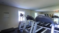 This single-story fitness room offers a practical and functional workout space designed to support cardiovascular exercise. The room features Precor-brand equipment, including at least two treadmills and an elliptical trainer, arranged to maximize the available floor space. A large wall mirror spans one side of the room, creating a sense of added depth while allowing users to monitor their form during workouts. The flooring is a dark, rubberized material well-suited for a fitness environment, providing durability and traction. Natural light enters through two windows fitted with horizontal blinds, keeping the space bright without harsh glare. The walls are painted in a neutral beige tone, contributing to a clean and understated aesthetic. Informational signage, including posted fitness center rules, is visible on the walls, indicating this is a shared or communal facility. The ceiling is standard height with a smooth white finish. Overall, the space is straightforward and purpose-driven, offering the essential equipment and environment needed for an effective cardio-focused workout routine.
