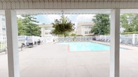 Located at 3500 Cypress Creek Dr in Florissant, Missouri, this community pool facility offers a thoughtfully designed single-level outdoor amenity space built for relaxation and socializing. The centerpiece is a generously sized swimming pool with crystal-clear blue water, surrounded by a classic brick-accented pool deck and enclosed by a white picket fence that lends a clean, welcoming aesthetic.

Adjacent to the pool is a spacious covered patio that provides a shaded retreat without sacrificing the open-air atmosphere. The patio is outfitted with ceiling fans to ensure comfortable airflow during warm Missouri summers, and hanging baskets of flowering plants add vibrant color throughout. Large multi-pane windows along the building facade allow natural light to flow through and provide clear sightlines to the pool area from inside.

The outdoor seating area features wicker sectional sofas and a coffee table arranged for comfortable conversation, making it an ideal gathering spot. A bold orange entry door serves as a distinctive architectural accent, offering direct access between the covered patio and the building interior. Lounge chairs are positioned poolside within the fenced enclosure for sunbathing. Mature trees visible beyond the fence provide a lush, green backdrop, enhancing the overall serene and inviting atmosphere of this community outdoor space.