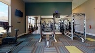 This fitness center is a comprehensively outfitted facility designed to support a wide variety of workout routines. The space is spread across a single floor and is organized into distinct zones, each dedicated to a different type of exercise. Large windows line the walls throughout, flooding the interior with natural light and offering views of the surrounding exterior — creating an open, energizing atmosphere enhanced by ceiling fans overhead.

The cardio zone features a row of treadmills, elliptical machines, and stair climbers positioned along the windowed walls, each equipped with individual monitors. A separate area houses stationary bikes and additional low-impact cardio equipment accompanied by a wall-mounted television. The strength training section is well-appointed with a multi-functional cable machine, a Smith machine with a bench press setup, power racks, and a dedicated free weights area stocked with dumbbells on tiered racks. A TRX suspension training rig and a hanging TKO heavy punching bag provide functional and combat fitness options.

Wall-to-wall mirrors in the strength area enhance the sense of space and allow users to monitor their form. The flooring throughout consists of colorful, multi-toned carpet tiles that add a modern, dynamic aesthetic to the space. The entrance features glass double doors, informational postings, and sanitation stations, ensuring a clean and welcoming environment from the moment of arrival.