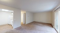 Located at 1716–1722 Bicentennial Way in North Providence, Rhode Island, this single-floor unit offers a practical and straightforward layout across several well-defined living spaces. The living room is generously sized, featuring warm neutral-toned carpeting, cream-colored walls, and a sliding glass door that floods the space with natural light and provides direct access to an outdoor area with a stone landscape backdrop. A wall-mounted air conditioning unit and baseboard heating ensure year-round comfort. The adjacent rooms, which serve as bedrooms or flexible living spaces, continue the carpeted flooring and are bright thanks to large windows overlooking the surrounding greenery. Each room benefits from baseboard heating and includes closet access. The kitchen is functional and well-appointed, featuring hardwood flooring, white cabinetry, dark granite-style laminate countertops, a stainless steel sink, a dishwasher, and a full-size refrigerator. The bathroom is clean and efficient, outfitted with a full soaking tub and shower combination framed by classic white tile surrounds and chrome fixtures. Throughout the unit, the light neutral palette and consistent finishes create a cohesive, move-in-ready environment suited for everyday living.