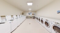 Located at 2401 6th St in Lake Charles, Louisiana, this shared laundry facility is a single-floor space designed to serve a high volume of users efficiently. The room is laid out in a clean, functional rectangular floor plan with white walls and light-colored flooring that contribute to a bright, sanitary atmosphere.

The facility is outfitted with two distinct rows of laundry machines lining opposing walls. One side features a lineup of top-loading washing machines, while the opposite wall houses a combination of front-loading washers and dryers. Together, the machines offer substantial capacity to accommodate multiple users simultaneously. Each unit is clearly numbered, making it easy for users to identify and manage their laundry.

Illumination is provided by flush-mounted overhead panel lights evenly distributed across the ceiling, ensuring the entire room is well-lit. A centrally located floor drain indicates practical design considerations for spills and maintenance.

Supporting amenities include stainless steel folding and sorting tables, as well as wheeled wire laundry carts for convenient transport. Informational signage is posted on the walls to guide users on proper machine operation and laundry procedures. The space is accessed through a solid entry door, with an additional glazed door providing secondary access. A bulletin board and fire safety equipment round out the practical features of this thoughtfully designed laundry facility.