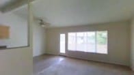 This single-floor space presents a straightforward and functional layout centered around a generously sized main living area. The room is finished with neutral-toned walls and light-colored carpeting, creating a clean and understated aesthetic throughout.

A dominant feature of the space is the expansive wall of windows paired with a sliding glass door, which spans much of the exterior-facing wall. This configuration allows an abundance of natural light to flood the interior, while also providing direct views of the surrounding outdoor greenery visible through the glass. The combination of the door and large windows creates a seamless visual connection between the interior and exterior.

The ceiling is smooth and painted white, contributing to a bright and open atmosphere. A ceiling fan is mounted centrally, offering air circulation throughout the room. A wall-mounted air conditioning unit is also installed, providing a dedicated climate control solution.

The overall ambiance of the space is simple and uncluttered, offering a neutral canvas that can be adapted to a variety of uses. The ample natural light and outdoor views lend the interior a welcoming and airy character.