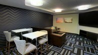 This single-floor space presents a thoughtfully designed lounge or gathering area that blends contemporary style with functional comfort. The room is anchored by a striking feature wall adorned with a bold chevron pattern in deep navy and charcoal tones, creating a dramatic focal point that sets the tone for the space's modern aesthetic. Complementing this, colorful wall art featuring vibrant cityscapes adds a lively, urban energy to the environment.

The flooring consists of patterned carpet tiles in a geometric arrangement of blues, grays, and whites, lending visual interest underfoot while defining the space with a professional yet inviting feel. Overhead, flush-mounted drum light fixtures provide clean, even illumination throughout.

Seating is thoughtfully arranged to accommodate a variety of uses. High-top tables paired with upholstered bar-height chairs occupy one section, ideal for casual meetings or independent work. A sectional seating area with a low central table offers a more relaxed lounge setting, suitable for collaborative gatherings or informal conversations. A built-in or freestanding divider helps subtly zone the space without fully enclosing it. The overall ambiance is modern and polished, making this space well-suited as a hospitality lounge, corporate common area, or hotel amenity space.