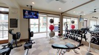 This modern fitness center is designed to offer a comprehensive workout experience within a thoughtfully designed, visually striking environment. The space is anchored by a distinctive scalloped-pattern flooring that flows throughout, lending a cohesive and stylish aesthetic to every area.

The cardio zone features a row of treadmills and elliptical machines positioned beneath soaring double-height ceilings with expansive windows that flood the room with natural light and offer pleasant outdoor views. Wall-mounted fans and an ornate pendant light add character to this open, airy section.

Adjacent to the cardio area is a dedicated free weights zone, outfitted with an extensive selection of dumbbells and kettlebells on tiered racks, along with multiple adjustable benches and medicine balls. A large floor-to-ceiling mirror visually expands the space and supports form-checking during workouts. A wall-mounted TV keeps members entertained, while large sliding glass doors offer direct views of an outdoor pool area.

The functional training zone includes a rowing machine, suspension training equipment, and plyo boxes, catering to a diverse range of fitness routines. The entryway connects these areas seamlessly, featuring exposed ductwork that adds an industrial-chic touch, a staircase with cable railings, and a striking blue geode art piece as a focal point. The overall ambiance is bright, modern, and energizing.