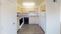 This single-story space presents a straightforward and functional layout, as illustrated by the overhead floor plan view. The interior is finished throughout in a neutral, cream-toned palette that lends a clean and cohesive aesthetic to every room.

The kitchen is equipped with light wood cabinetry offering ample storage, a double-basin sink, an electric range, a dishwasher, and a full-size refrigerator. Dark hardwood-style flooring grounds the space, and overhead fluorescent lighting ensures excellent visibility during meal preparation.

The living area is generously proportioned, featuring plush carpeting, vertical blinds along a large sliding glass door, and an additional window that allows natural light to fill the room. A separate entry door provides convenient access.

Multiple additional rooms—suitable for bedrooms or flexible use—are carpeted and feature individual windows with blinds, providing natural light and a comfortable atmosphere. One room includes a dedicated utility hookup, adding practical functionality.

The bathroom is well-appointed with a full tub-and-shower combination, white tile surround, a toilet, and a vanity with countertop storage. Wood-style vinyl flooring completes the space with a clean, durable finish.

Overall, the space maintains a simple, livable character with consistent finishes throughout.
