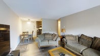 This single-floor space offers a comfortable and functional open-concept layout that seamlessly connects the main living, dining, and kitchen areas. The living room serves as the heart of the home, featuring warm hardwood-style flooring, neutral gray walls, and a cozy seating arrangement with two patterned sofas centered around a wood and glass coffee table. A floor lamp provides warm ambient lighting, complementing the inviting atmosphere. The dining area is positioned adjacent to the living room and is furnished with a wooden table and matching chairs, ideal for everyday meals. A ceiling fan overhead adds both style and practicality to this zone. The kitchen is visible from the open living and dining space, featuring dark cabinetry, stainless steel appliances including a microwave and range, and efficient use of the available space. The consistent hardwood-style flooring flows throughout the main areas, creating a cohesive and unified aesthetic. The neutral color palette, warm lighting, and open arrangement make this a welcoming and practical single-floor residence well-suited for comfortable everyday living.