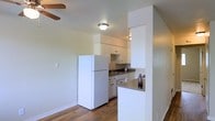This single-floor space offers a clean, functional layout with an open-concept design that connects the kitchen and living area seamlessly. The kitchen is outfitted with white cabinetry, a dark countertop, a sink, and full-sized appliances including a refrigerator. A partial wall with a breakfast bar counter serves as a natural divider between the kitchen and the adjoining living space, adding both definition and convenience to the layout. The flooring throughout features warm hardwood-style planks that contribute to a cohesive and inviting atmosphere. Neutral-toned walls keep the interior feeling bright and airy, while flush-mount ceiling lighting ensures consistent illumination. A ceiling fan is visible in the living area, providing both comfort and airflow. The space benefits from a straightforward, efficient design that prioritizes practicality while maintaining a comfortable and welcoming ambiance. Overall, the layout makes good use of its footprint, offering an approachable and livable environment with thoughtful details throughout.