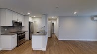 This single-floor space showcases a thoughtfully designed open-concept layout that seamlessly connects the kitchen and living areas. The kitchen is a standout feature, anchored by a central island with a dark countertop and an undermount sink, providing both additional prep space and a natural gathering point. White cabinetry lines the perimeter walls, offering generous storage, while stainless steel appliances — including a French door refrigerator, range, and microwave — lend a sleek, contemporary feel. Recessed lighting is installed throughout, ensuring consistent, clean illumination across the space. Warm hardwood-style flooring flows continuously from the kitchen into the open living area, creating a cohesive and inviting atmosphere. The soft, light-gray walls complement the neutral palette and contribute to a bright, airy environment. A hallway visible from the main living area leads to additional rooms, suggesting a practical and well-organized floor plan. The overall design balances modern aesthetics with functional living, making efficient use of the available space on a single level.