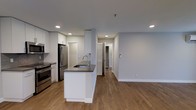 This single-floor space showcases a thoughtfully designed open-concept layout that seamlessly connects the kitchen and living areas. The kitchen is a standout feature, anchored by a central island with a dark countertop and an undermount sink, providing both additional prep space and a natural gathering point. White cabinetry lines the perimeter walls, offering generous storage, while stainless steel appliances — including a French door refrigerator, range, and microwave — lend a sleek, contemporary feel. Recessed lighting is installed throughout, ensuring consistent, clean illumination across the space. Warm hardwood-style flooring flows continuously from the kitchen into the open living area, creating a cohesive and inviting atmosphere. The soft, light-gray walls complement the neutral palette and contribute to a bright, airy environment. A hallway visible from the main living area leads to additional rooms, suggesting a practical and well-organized floor plan. The overall design balances modern aesthetics with functional living, making efficient use of the available space on a single level.