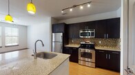 This well-appointed single-floor residence offers a thoughtfully designed open-concept layout that seamlessly connects the kitchen, dining, and living areas. The kitchen is a standout feature, equipped with a full suite of stainless steel appliances including a refrigerator, electric range, and built-in microwave. Rich, dark espresso cabinetry extends floor to ceiling, complemented by a mosaic tile backsplash and light granite countertops. A generously sized center island with an undermount sink and pendant lighting serves as both a functional workspace and a natural gathering point. Warm hardwood-style flooring flows consistently throughout the main living areas, adding a sense of continuity and warmth. The living area benefits from multiple windows and a glass-paneled door that invite abundant natural light, while a ceiling fan adds comfort and airflow. Track lighting in the kitchen provides focused, adjustable illumination. The bathroom features matching granite countertops, a dark-framed vanity mirror, and a full tub-shower combination with tile surround. Additional rooms include a carpeted bedroom and a flexible secondary room with hardwood-style flooring, suitable for a bedroom, office, or den. The space presents a clean, contemporary aesthetic throughout.