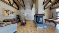 This single-floor space makes a powerful architectural statement with its dramatic vaulted ceilings adorned by a series of exposed, dark-stained wood beam trusses that arc gracefully overhead, lending the interior a warm, lodge-inspired character. The expansive main living area is anchored by a standout three-sided brick fireplace at its center, featuring a sleek black surround, a reclaimed wood mantel, and a mounted flat-screen television above — serving as both a functional and visual focal point that can be appreciated from multiple vantage points throughout the room.

Wide-plank hardwood flooring in a natural, light-toned finish runs throughout the space, complementing the rustic aesthetic while keeping the interior feeling bright and open. The living area is generously proportioned, comfortably accommodating multiple seating arrangements centered around a woven area rug and a solid wood coffee table. Recessed lighting supplements the natural light that filters in through transom windows and a set of tall, glass-paneled entry doors near the front of the space.

Toward the entrance, a billiards table adds a recreational element, reinforcing the space's communal and entertaining character. The overall ambiance balances rustic warmth with open, airy grandeur — making it a welcoming and versatile gathering space.