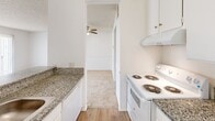 Located on the second floor at 20939 Parthenia Street in Canoga Park, California, Unit 218 is a one-bedroom residence offering a well-organized single-level layout. The galley-style kitchen is a functional highlight, featuring granite-patterned countertops, abundant white cabinetry extending to the ceiling, a double stainless steel sink, and a full suite of white appliances including a refrigerator and electric range with overhead ventilation. Hardwood-style flooring runs throughout the kitchen, lending a clean, cohesive look. The kitchen opens directly into the living area, where carpet flooring and a ceiling fan contribute to a comfortable, casual atmosphere. A large window fitted with vertical blinds allows natural light into the living space while providing views of the surrounding neighborhood. The bedroom is a generously proportioned room featuring wall-to-wall carpet, a textured ceiling, and a dedicated HVAC vent for climate control. An adjacent walk-in closet provides substantial storage capacity, with wrap-around hanging rods and carpet flooring. The overall design of the unit is clean and neutral throughout, with white walls creating a bright, open feel well-suited for everyday living in the Canoga Park community.