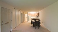 This single-floor residential unit offers a clean, functional layout with an open-plan design that seamlessly connects the living, dining, and kitchen areas. The space is finished in warm neutral tones, with light-colored walls and plush carpeted flooring that contribute to a comfortable, inviting atmosphere throughout.

The dining area is positioned at the heart of the unit, comfortably accommodating a four-seat dining table with wicker-style chairs — ideal for everyday meals or casual entertaining. A set of full-height sliding closet doors lines one wall, providing generous built-in storage without compromising the openness of the space.

The kitchen is situated at the rear of the main living area and features light wood cabinetry, ample counter space, and essential appliances including a full-size refrigerator and stovetop. The layout is practical and efficient, making good use of the available footprint.

A hallway leads further into the unit, connecting additional rooms and suggesting a well-organized floor plan. Overhead lighting illuminates the kitchen area, while the overall layout benefits from a sense of openness and flow. The consistent neutral palette and simple finishes throughout give the space a clean, move-in-ready character suitable for a range of lifestyles.