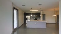This single-floor space showcases a contemporary open-concept design that seamlessly integrates the living and kitchen areas into one cohesive environment. The kitchen is a standout feature, equipped with stainless steel appliances including a French-door refrigerator, electric range, and built-in microwave. Dark espresso cabinetry extends to the ceiling, providing ample storage while creating a bold contrast against the light-toned countertops. A central island with a white finish and integrated sink serves as both a functional workspace and a natural dividing point between the kitchen and living area. Pendant lighting fixtures hang above the island, complementing the overhead flush lighting. The flooring throughout is a smooth, polished concrete, lending an industrial yet refined aesthetic to the space. A large sliding glass door with slatted blinds along one wall floods the interior with natural light and hints at outdoor access. The neutral wall tones and clean lines throughout contribute to a bright, minimalist atmosphere that feels both spacious and functional.