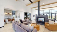 Located at 3851 N Traverse Mountain Blvd in Lehi, Utah, this single-floor community space offers a well-appointed and thoughtfully designed gathering environment. The expansive living room is anchored by soaring ceilings with warm wood beam accents, polished concrete flooring, and floor-to-ceiling steel-framed glass walls that flood the interior with natural light and open directly to the outdoors. A central fireplace serves as a striking focal point, complemented by a large communal dining table, eclectic seating arrangements, and a ceiling fan that enhances the airy atmosphere. A separate lounge area features sage green walls, a tan leather sectional, and a mounted TV, offering a more intimate retreat within the space. The kitchen is sleek and modern, showcasing white subway tile backsplash, dark countertops, white cabinetry, stainless steel appliances, and a stunning marble-topped island with leather bar seating. Brass sconce lighting and open shelving add refined character. The outdoor patio extends the living space with a steel pergola providing partial shade over dining tables and chairs, alongside outdoor grills and open lawn space, all set against views of the surrounding Traverse Mountain community.