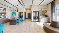 This contemporary lobby space offers a thoughtfully curated environment that blends professional functionality with inviting design. The open, single-floor layout is anchored by large-format light tile flooring that reflects natural light streaming in through expansive floor-to-ceiling windows, providing outdoor views of the surrounding grounds. Soaring ceilings with decorative wood beam accents lend architectural character and a sense of openness throughout the space.

A dedicated workstation area features a substantial dark wood table paired with vibrant teal velvet chairs and suspended black-and-gold pendant lighting, creating a striking focal point. Framed black-and-white photography and curated shelving with decorative accessories add a polished, gallery-like quality to the walls. Adjacent to the workstation is a built-in service counter finished in warm wood tones with a chevron-patterned backsplash and granite countertop, ideal for hospitality or administrative use.

Glass-paneled interior doors and partitions connect the main lobby to additional meeting or conference areas, maintaining visual openness while defining separate zones. Near the entrance, a curved, modern lounge seat offers a welcoming spot for visitors. Dark-framed windows and doors throughout provide clean contrast against the neutral palette. The overall ambiance is contemporary, warm, and professionally appointed.