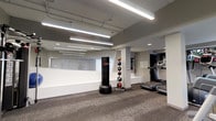 This fitness facility offers a well-equipped, single-floor exercise space designed to support a variety of workout routines. The interior features a neutral color palette of whites and grays, complemented by carpet flooring that adds comfort underfoot during training sessions. Bright fluorescent strip lighting runs along the exposed ceiling, ensuring the space is thoroughly illuminated throughout.

The layout is open and organized, with distinct zones for different types of exercise. A freestanding heavy punching bag anchors one area, providing a dedicated space for combat-style training. Adjacent to it, a tiered medicine ball rack holds an assortment of weighted balls, catering to functional and strength training exercises. On the opposite side, multiple treadmills are positioned along the wall, offering cardio options for users.

A large mirror spans one wall, enhancing the sense of openness and allowing users to monitor their form during workouts. Structural columns are integrated naturally into the design without interrupting the flow of the space. The overall ambiance is clean, modern, and practical, making it a functional environment suited for both cardio and strength-based fitness activities.