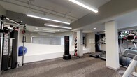 This fitness facility offers a well-equipped, single-floor exercise space designed to support a variety of workout routines. The interior features a neutral color palette of whites and grays, complemented by carpet flooring that adds comfort underfoot during training sessions. Bright fluorescent strip lighting runs along the exposed ceiling, ensuring the space is thoroughly illuminated throughout.

The layout is open and organized, with distinct zones for different types of exercise. A freestanding heavy punching bag anchors one area, providing a dedicated space for combat-style training. Adjacent to it, a tiered medicine ball rack holds an assortment of weighted balls, catering to functional and strength training exercises. On the opposite side, multiple treadmills are positioned along the wall, offering cardio options for users.

A large mirror spans one wall, enhancing the sense of openness and allowing users to monitor their form during workouts. Structural columns are integrated naturally into the design without interrupting the flow of the space. The overall ambiance is clean, modern, and practical, making it a functional environment suited for both cardio and strength-based fitness activities.