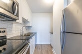 Located at 21 King Street in downtown London, Ontario, this two-storey, 2-bedroom, 2-bathroom unit offers a practical and well-appointed layout spread across two levels. The main floor is a compact, functional entry level featuring light wood-style flooring, a private entry door with windows that bring in natural light, and a baseboard heating system. The kitchen is outfitted with white shaker-style cabinetry, dark countertops, a subway tile backsplash, a double stainless steel sink, and stainless steel appliances including a range and over-the-range microwave. A half bathroom on this level is finished in clean white tile with a wall-mounted sink and modern fixtures. A staircase with white railings connects the two floors. The upper level houses the primary and secondary bedrooms, both featuring light wood flooring, closets, and baseboard or wall-mounted heating units. The primary bedroom is a comfortable size with a sliding closet. The full bathroom on this floor includes a tiled tub-shower combo with accent tile, a vanity, and stone-look tile flooring. The spacious living area on the upper level is highlighted by large floor-to-ceiling sliding glass doors opening to a balcony with open views of the surrounding treetops and cityscape.