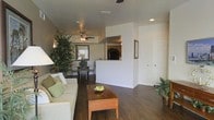 This single-floor residence offers a thoughtfully designed open-concept layout that connects the living, dining, and kitchen areas in one cohesive flow. The living room is anchored by a comfortable cream sofa adorned with green accent pillows and a warm wood coffee table, creating an inviting and functional gathering space. Dark hardwood flooring runs throughout, lending a rich contrast to the crisp white walls and adding a sense of warmth and continuity across the space. A ceiling fan with integrated lighting serves both the living and dining zones, promoting comfortable airflow throughout. The dining area is positioned near a window that invites natural light, accompanied by a tall, lush indoor tree that adds a vibrant organic element to the interior. A half-wall partition elegantly separates the kitchen from the living area while maintaining an open feel, with a pass-through window allowing visual connectivity between spaces. The kitchen features a clean white countertop visible from the living area. Recessed lighting and neutral wall tones enhance the brightness of the space, while indoor plants placed strategically throughout contribute to a fresh, welcoming ambiance. The layout is both practical and aesthetically balanced, making it well-suited for everyday living and entertaining alike.