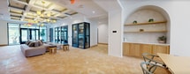 Located at 85 Livingston Ave in Roseland, NJ, this 1,170-square-foot single-floor amenity lobby offers a thoughtfully designed communal space that blends functionality with refined aesthetics. The space is characterized by warm terracotta herringbone tile flooring, crisp white walls, and distinctive coffered ceilings with gold accents, creating an upscale yet welcoming atmosphere throughout.

The open-plan layout is divided into several functional zones. A lounge area features comfortable sectional sofas, marble-topped accent tables, and cylindrical stools, ideal for relaxed gatherings. Adjacent to this, elegant arched architectural details frame a dining area furnished with a leather sofa and bistro-style chairs, beneath a striking brass chandelier.

A standout feature is the row of glass-enclosed private work pods with dark-framed doors, each equipped with a desk and task chair, providing quiet, focused workspaces within the communal environment. The mailroom area is outfitted with an extensive wall of built-in residential mailboxes framed by warm wood cabinetry, accompanied by additional lounge seating. Large floor-to-ceiling windows and glass entry doors flood the space with natural light, offering views of lush greenery outside. Recessed lighting and pendant chandeliers with frosted amber shades complete the polished, contemporary design.