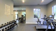 Located at 3590 Mary Adele Ave in Charleston, SC, this fitness center offers a well-appointed, single-floor exercise facility designed to support a comprehensive workout experience. The space is finished with durable, textured flooring suited for high-traffic athletic use, and the neutral gray walls create a clean, focused environment. Recessed lighting throughout ensures consistent illumination across the workout areas, while multiple windows allow natural light to filter in and provide views of the surrounding greenery, contributing to an open and energizing atmosphere. The facility is divided into distinct workout zones. The free weights area features a full dumbbell rack with a wide range of weight increments, ideal for strength training routines. The cardio section includes a stair climber and additional cardio equipment positioned near the windows. A dedicated strength training zone houses cable and pulley machines, a multi-function weight station, and additional resistance equipment, offering users a variety of exercise options. A large wall mirror in the strength area enhances the sense of space and allows for proper form monitoring during workouts. The layout provides ample room to move between stations comfortably. Overall, this fitness center is a practical, well-equipped space suited for residents seeking convenient on-site exercise amenities.