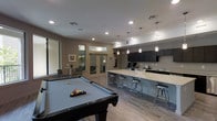 This single-floor amenity space is thoughtfully designed to blend social entertainment with functional gathering areas. The open-concept layout centers around a full-sized pool table positioned on wood-look plank flooring, creating an inviting recreational focal point. Adjacent to the game area, a spacious kitchen island topped with light quartz countertops is lined with modern black metal bar stools, offering ample seating for socializing. A series of elegant glass pendant lights hang above the island, complementing the recessed ceiling lighting throughout. The kitchen is well-appointed with dark cabinetry, a contrasting light backsplash, and a full sink setup, making it ideal for catering or casual use. A glass-paneled door leads to a secondary room visible in the background, suggesting additional flexible space for meetings or private gatherings. Neutral taupe walls and consistent flooring tie the areas together, giving the space a cohesive, contemporary feel. The combination of a game area, communal kitchen, and lounge zones makes this a versatile and well-equipped shared amenity space suited for a variety of group activities and events.