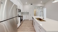 This single-floor unit presents a clean, modern aesthetic defined by a bright white color palette, light wood-look luxury vinyl plank flooring, and an open layout that promotes a sense of spaciousness. The open-concept kitchen and living area form the heart of the unit, where the kitchen stands out with tall white shaker cabinets, a subway tile backsplash, stainless steel appliances including a refrigerator, microwave, and range, and a generous island featuring a deep undermount sink with a commercial-style faucet and pendant lighting above. Recessed lighting throughout keeps the kitchen and living areas well-illuminated. The living space connects to a sliding glass door that opens to an outdoor area, allowing natural light to pour in and providing outdoor views. The bedroom is carpeted for comfort and features a large window fitted with blinds, a ceiling vent, and access to a spacious walk-in closet equipped with a full-width hanging rod and shelving. The bathroom is finished with light wood-look flooring, a wide white vanity with ample drawer storage, quartz countertops, a large mirror, and a full soaking tub and shower combination. The overall ambiance is fresh, contemporary, and move-in ready.