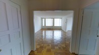 This single-floor space offers a clean, well-maintained interior defined by timeless design elements and a functional layout. The standout feature of the main living area is the beautifully patterned parquet hardwood flooring, finished to a high sheen that reflects the generous natural light pouring in through a row of large windows along the far wall. These windows create a bright, airy atmosphere throughout the space.

The living area is accessed through a wide, open doorway that provides a clear sightline across the room, enhancing the sense of openness and flow. The neutral white walls keep the interior feeling fresh and versatile, offering a blank canvas suitable for a variety of uses and furnishing styles.

The layout appears thoughtfully arranged, with additional doorways visible from the main living area suggesting connected rooms that contribute to a practical and cohesive floor plan. The baseboard heating visible along the windows indicates year-round comfort within the space.

Overall, the space combines classic architectural details — most notably the parquet flooring — with a bright, minimalist aesthetic that makes it feel both inviting and adaptable. The abundance of natural light and open proportions of the main room are among its most defining and desirable characteristics.