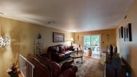 Located at 1211 White Pine Cir in Lawrence Township, NJ, this single-floor home offers a well-organized layout encompassing one bedroom, one bathroom, two living areas, a dining room, and a kitchen. The main living room features warm golden-toned walls, hardwood flooring, and a standout sliding glass door that floods the space with natural light and opens to a deck overlooking lush greenery — an inviting indoor-outdoor connection. A secondary living area, painted in cool slate blue with decorative starburst wall accents, provides a quieter retreat. The dining room is finished in cheerful yellow tones, centered around a classic chandelier and a round wood dining table — a welcoming space for meals. The kitchen is equipped with rich dark wood cabinetry, a mosaic tile backsplash, and ample counter space in a practical corner layout. The spacious primary bedroom carries warm amber walls accented with a decorative wallpaper border, offering a cozy and private sanctuary. The bathroom is cleanly finished with neutral tile wainscoting, a vanity with a framed mirror, and tasteful wall art. Throughout the home, wood-toned flooring and warm color palettes create a cohesive, comfortable ambiance.