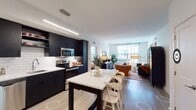 This well-appointed single-floor residence combines modern design with functional living across a thoughtfully arranged floor plan. The open-concept kitchen is a standout feature, showcasing bold navy cabinetry, white subway tile backsplash, stainless steel appliances, and a central island with a white countertop that doubles as a dining area. Light wood-tone flooring runs throughout the main living spaces, enhancing the bright, airy atmosphere created by large windows that allow ample natural light.

The living room offers a comfortable and inviting layout with generous window views of the surrounding neighborhood. The space flows seamlessly from the kitchen, creating an ideal setting for both relaxing and entertaining.

Two bedrooms provide distinct retreats. The primary bedroom features a calming accent wall, plush carpeting, a dedicated workspace, and access to a spacious walk-in closet equipped with wire shelving for ample storage. The secondary bedroom is equally well-lit, with a large window and soft carpeting.

The bathroom is finished with dark tile flooring, a navy vanity with a white countertop, a large framed mirror, and clean white walls, maintaining the cohesive modern aesthetic found throughout the residence. Overall, the space balances contemporary style with practical, comfortable living.