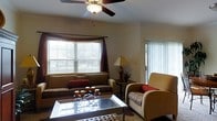 This single-floor living space centers around a comfortable and well-appointed living room designed for relaxed everyday use. The room is illuminated by abundant natural light streaming through a wide horizontal window fitted with white blinds and accented by rich, dark burgundy curtains that add a warm, decorative contrast to the neutral walls. A sliding glass door on the adjacent wall introduces additional light and provides outdoor access, enhancing the open feel of the space.

A ceiling fan with an integrated light fixture is centrally positioned overhead, offering both practical air circulation and supplemental lighting. The living area is furnished with a plush, earth-toned sofa accented by red throw pillows, complemented by a cushioned armchair in warm gold tones. A glass-top coffee table with decorative metalwork sits at the center of the seating arrangement atop a patterned area rug, adding visual interest to the room.

A traditional table lamp and a small decorative plant on a side table complete the warm, layered ambiance. The overall aesthetic is casual and inviting, with a cohesive color palette of warm neutrals, burgundy, and gold tones throughout.