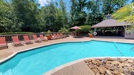 Located at 711 Harbour Dr in Ridgeland, Mississippi, this outdoor amenity space offers a well-appointed poolside retreat designed for relaxation and leisure. The centerpiece is a generously sized freeform swimming pool with crystal-clear blue water, accented by a decorative natural stone border along one edge that adds a resort-like aesthetic. Surrounding the pool is a spacious textured concrete sundeck finished in a warm terracotta tone, providing ample room for lounging and socializing. A collection of red sling-back lounge chairs and side tables are arranged along the deck, offering comfortable spots to enjoy the outdoors. A large patio umbrella provides additional shade for those seeking relief from the sun. At one end of the deck, a covered wooden gazebo serves as a shaded gathering area, equipped with seating and what appears to be informational or safety signage. The entire space is enveloped by tall, mature deciduous trees that create a lush, private canopy, enhancing the sense of seclusion and natural beauty. The single-level layout ensures easy accessibility throughout the area, making it a functional and inviting outdoor amenity for residents or guests.