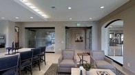 This polished single-floor space presents a refined and professional atmosphere designed for comfort and functionality. The main lounge area is anchored by a well-appointed seating arrangement featuring tufted upholstered armchairs in neutral tones, complemented by a decorative bistro-style side table and a patterned area rug that adds visual warmth to the light-tiled flooring. A dining or meeting area with dark chairs is positioned nearby, offering a versatile setting for gatherings or collaborative work.

The architectural design highlights include a tiered ceiling with recessed lighting accented by distinctive blue LED strips, which lend a contemporary and upscale character to the space. Full-height glass partition walls with dark framing define adjacent rooms while maintaining an open, transparent feel throughout, allowing light to flow freely and preserving sightlines across the floor plan.

The neutral warm-gray wall palette and clean architectural lines create a calm, sophisticated backdrop. Thoughtful decorative touches, including wall art and subtle greenery, add personality without overwhelming the space. The overall ambiance balances modern design sensibility with welcoming comfort, making it well-suited for use as a residential amenity lounge, boutique office reception, or upscale collaborative gathering area.