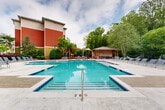 Located at 1321 Lovvorn Rd in Carrollton, Georgia, this outdoor amenity space centers around a generously sized swimming pool with crystal-clear turquoise water. The pool features a multi-section layout with varying depths, clearly marked entry points, and blue tile accents along the waterline, offering a resort-like experience for residents and guests.

Surrounding the pool is a well-appointed sundeck finished in light-toned concrete and patterned paver surfaces, lined with rows of adjustable chaise lounge chairs ideal for relaxing in the Georgia sun. A dedicated lounge zone near the tree line includes hammocks strung between posts and additional seating beneath a rustic wooden pergola, creating a shaded retreat with a natural, park-like atmosphere.

A poolside clubhouse structure sits adjacent to the pool area, featuring large glass doors, warm wood detailing, and a covered porch with outdoor seating. A gazebo-style pavilion with additional table-and-chair seating provides a shaded gathering spot nearby, complete with umbrella tables for casual outdoor dining or socializing.

The entire area is enveloped by mature trees that provide privacy, natural shade, and a serene backdrop. Decorative lamp posts are distributed throughout the space, ensuring the area is well-lit during evening hours. The combination of thoughtful landscaping, multi-use amenity zones, and a striking pool makes this outdoor space a standout feature of the property.