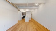 Located in the heart of Lancaster, PA, this one-bedroom, three-bathroom loft blends industrial character with modern finishes across a thoughtfully designed single-floor layout. The open-concept living and kitchen area immediately impresses with soaring ceilings, exposed metal ductwork, corrugated steel ceiling panels, and warm hardwood flooring that runs throughout the main living space. A central kitchen island anchors the space, complemented by a well-appointed kitchen featuring taupe shaker-style cabinetry, quartz countertops, stainless steel appliances, and hardwood flooring. Natural light filters in through large windows, offering views of the surrounding brick architecture of downtown Lancaster. A hallway leads to the private bedroom suite, which is carpeted for comfort and includes a window framing the urban streetscape, generous closet space with built-in rods, and direct access to the bathrooms. The bathrooms are finished with neutral tile flooring, dark wood vanities, LED-lit mirrors, and glass-enclosed walk-in showers with tile surround. Additional hallways provide practical utility space, completing this well-organized urban residence.