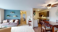 This single-floor residence offers a thoughtfully arranged layout with three bedrooms, one bathroom, a living room, a combined dining room and kitchen, and a central hallway connecting the spaces. The living room is a bright, welcoming area featuring carpet flooring, a teal accent wall with floating shelves, and a sliding glass door that fills the room with natural light and provides outdoor access. Adjacent to the living room, the dining area features hardwood-style flooring and a decorative round mirror, creating an intimate setting for meals. The galley-style kitchen is equipped with dark wood cabinetry, granite-style countertops, a coil-burner range, refrigerator, and a stainless steel sink, offering efficient use of space. The primary bedroom is generously sized, featuring carpet flooring, a large double closet with bi-fold doors, and warm neutral tones. The second bedroom includes a full-width window with vertical blinds and its own closet. The full bathroom is finished with tile flooring, a granite-style vanity countertop, dark wood cabinetry, a large mirror, and a tub-shower combination. Throughout the residence, the neutral color palette and consistent finishes create a cohesive and comfortable living environment.