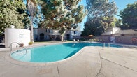 Located at 750 Mobil Ave in Camarillo, California, this outdoor pool area offers a well-appointed communal retreat designed for relaxation and recreation. The centerpiece is a generously sized freeform swimming pool with clear, blue water, framed by a wide concrete sundeck that provides ample space for lounging and movement around the pool perimeter. The pool features built-in entry steps and metal handrails for safe and easy access.

Surrounding the pool, a mature landscape of tall palm trees and large evergreen and deciduous trees creates a lush, shaded canopy that adds privacy and a resort-like ambiance to the space. The natural greenery softens the hardscape and provides welcome shade during warm Southern California days. Outdoor seating areas are available on the deck, including patio tables with umbrellas and lounge chairs, offering comfortable spaces for poolside gatherings.

The area is enclosed by a metal security fence with gated access, ensuring safety and controlled entry. An adjacent low-rise residential building is visible in the background, reflecting the community-oriented nature of this amenity space. Safety equipment, including life rings, is stationed poolside in compliance with standard pool safety requirements. The overall setting benefits from abundant natural sunlight and a tranquil, tree-lined atmosphere characteristic of the Camarillo, CA region.