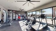 Located in Tolleson, Arizona, this dedicated fitness center is a well-appointed exercise facility designed to support a comprehensive workout experience. The space features a dark, rubberized floor that provides both durability and comfort underfoot, complemented by a crisp white ceiling outfitted with large ceiling fans to maintain airflow during intense sessions.

The facility is equipped with a diverse range of cardio machines, including treadmills, ellipticals, and stationary bikes from Spirit and other brands. A tiered dumbbell rack offers a full range of free weights, while additional strength equipment such as a cable/weight machine and rowing machine round out the training options. Exercise balls and medicine balls are also available for functional fitness training.

One of the standout design elements is the dramatic large-scale athletic mural painted across a dark accent wall, depicting a muscular athlete in motion — creating a motivating, energetic ambiance. Double glass doors open to the exterior, flooding the space with natural light, while a full wall of floor-to-ceiling windows along one side offers expansive views of the outdoor pool area and palm trees beyond.

High windows on additional walls bring in further natural light, keeping the space feeling bright and open despite its focused footprint. The overall aesthetic is sleek and contemporary, making this fitness center a functional and visually compelling space for residents in Tolleson, AZ.