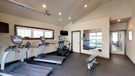 Located at 1605 North River Ridge Boulevard in Spokane, Washington, this single-floor fitness center is thoughtfully designed to support a wide range of workout routines. The space features soaring vaulted ceilings with recessed lighting throughout, creating an open and energizing atmosphere. Durable rubber flooring spans the entire area, providing a safe and functional surface for all types of exercise.

The cardio zone is well-equipped with multiple treadmills, elliptical trainers, and a rowing machine, complemented by wall-mounted flat-screen televisions for entertainment during workouts. A full-length mirror along one wall visually expands the space and allows exercisers to monitor their form. A dedicated strength training area includes a power rack with TRX suspension trainers, a dumbbell rack, medicine balls, and a flat utility bench, offering everything needed for a comprehensive strength session.

Natural light fills the space through several framed windows and a large glass overhead garage door, which can open to seamlessly connect the interior to the outdoor patio area. The warm, neutral-toned walls and dark wood trim details lend the space a clean, polished aesthetic. Additional amenities include a restroom and convenient access doors, making this fitness center a well-rounded and inviting facility for daily use.