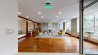 This single-floor amenity space is thoughtfully designed to accommodate a variety of functions, combining a welcoming lobby entrance, relaxed lounge areas, casual dining, and semi-private workspaces within one cohesive layout. Rich hardwood flooring extends throughout the majority of the space, providing warmth and continuity between zones.

The main lounge area serves as the central focal point, anchored by a large statement pendant light with a gold interior finish. Comfortable seating—including sculptural wingback chairs in olive green and a neutral upholstered sofa—surrounds clustered white pedestal coffee tables atop a soft area rug. Framed artwork and large windows allow natural light to fill the space and offer views to the outdoors.

Adjacent to the lounge, a casual dining area features built-in bench seating with patterned wallcovering panels framed in warm wood, paired with clean white tables and green accent chairs. A refreshment counter runs along one wall.

A secondary lounge zone features rich walnut wall paneling, a wall-mounted television, and a tray ceiling detail, furnished with a bold orange accent chair and neutral sectional sofa with patterned cushions. The glass-fronted entry vestibule completes the layout, providing a polished, well-lit arrival experience.