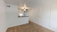 This single-story unit in Indianapolis, Indiana, offers a practical and efficiently organized layout across its one floor. The interior is finished with neutral white walls and textured ceilings throughout, creating a clean, consistent aesthetic. Carpeting covers the living room and bedroom, adding warmth underfoot, while the kitchen features wood-look vinyl plank flooring for easy maintenance.

The living room is an open, simply appointed space with a large window that draws in natural light and offers views of surrounding greenery. A ceiling fan is positioned near the pass-through counter that connects to the kitchen, promoting airflow between the two areas. The kitchen is galley-style, lined with white cabinetry, dark countertops, subway tile backsplash, and includes a refrigerator, electric range, and double-basin sink — all white appliances that complement the cabinetry.

The bedroom is a generously proportioned room with a window providing natural light and a baseboard heater along the wall. White panel walls add subtle texture, and a dedicated closet provides storage. The overall ambiance of the unit is understated and functional, with a bright, neutral palette throughout.