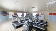 Located at 1800 Links Boulevard in Tuscaloosa, Alabama, this fitness center offers a dedicated, single-floor workout facility designed to support an active lifestyle. The space is outfitted with a robust selection of cardio equipment, including multiple treadmills and elliptical machines arranged to maximize floor space and ease of movement between stations. A mounted flat-screen television is positioned for convenient viewing during workouts, adding to the overall exercise experience. Natural light filters in through several windows along the walls, creating a bright and energizing atmosphere. A ceiling fan provides additional air circulation throughout the room, enhancing comfort during intense sessions. The walls feature a two-tone color scheme with warm beige tones accented by bold red panels, giving the space a motivating and purposeful aesthetic. An inspirational wall decal reading "Hard work keeps the dreams alive" reinforces the fitness-focused environment. The flooring is designed to support heavy equipment while remaining practical for varied workout routines. Overall, the facility presents a clean, functional, and well-maintained environment suited for cardiovascular training and general fitness activity.